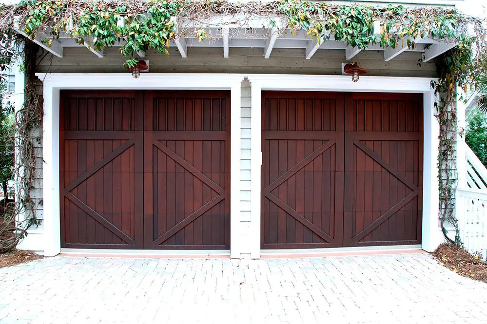 Custom garage door made from high-quality materials, featuring a unique design with decorative windows and hardware. The door is set in a well-maintained area with natural light casting shadows, enhancing its texture and color. Ideal for homeowners and designers looking for bespoke garage solutions.